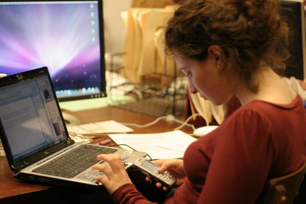 A girl navigates her phone while using her laptop while studying at home